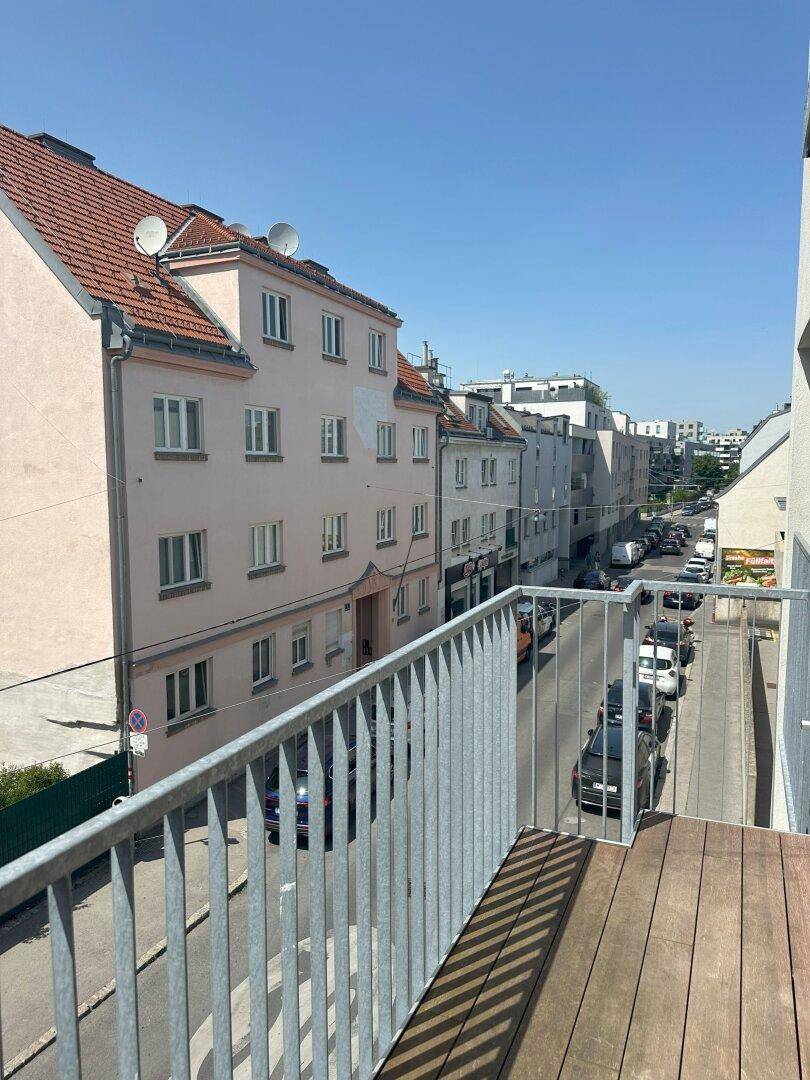 Blick vom Balkon auf eine belebte Straße mit angrenzenden Wohngebäuden unter blauem Himmel.