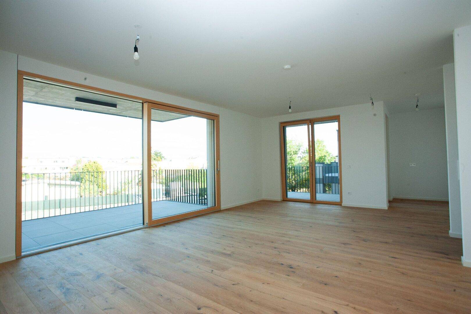 Geräumiges, leeres Wohnzimmer mit Holzboden und großen Schiebetüren, die auf einen Balkon führen.