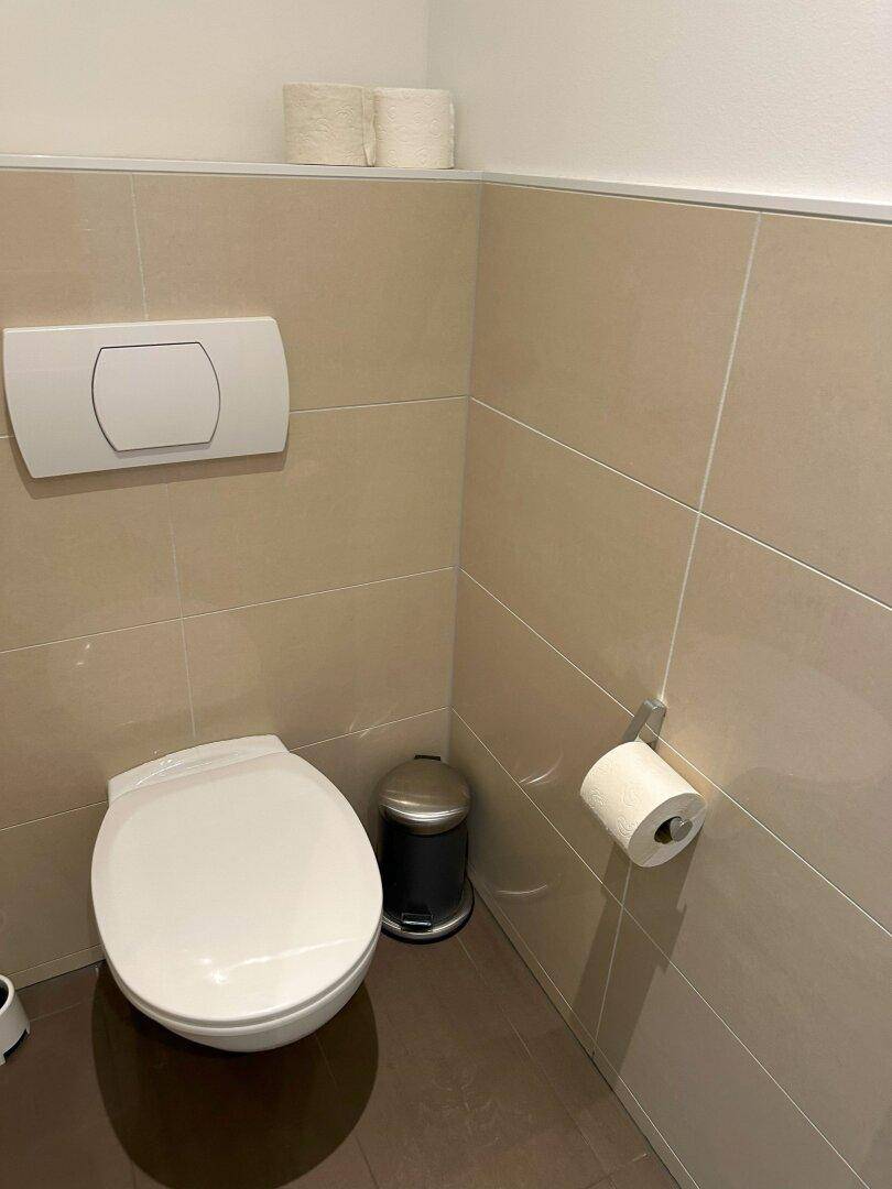 Sauberer Toilettenbereich mit wandhängendem WC, Spülkasten und einem kleinen Abfalleimer.