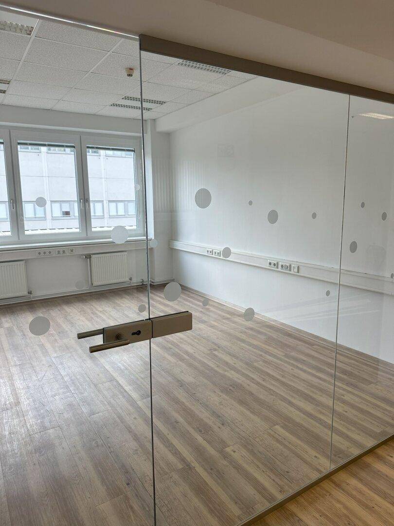 Leeres Büro mit großen Fenstern, Heizkörpern und einer modernen Glastür mit Punktmuster.