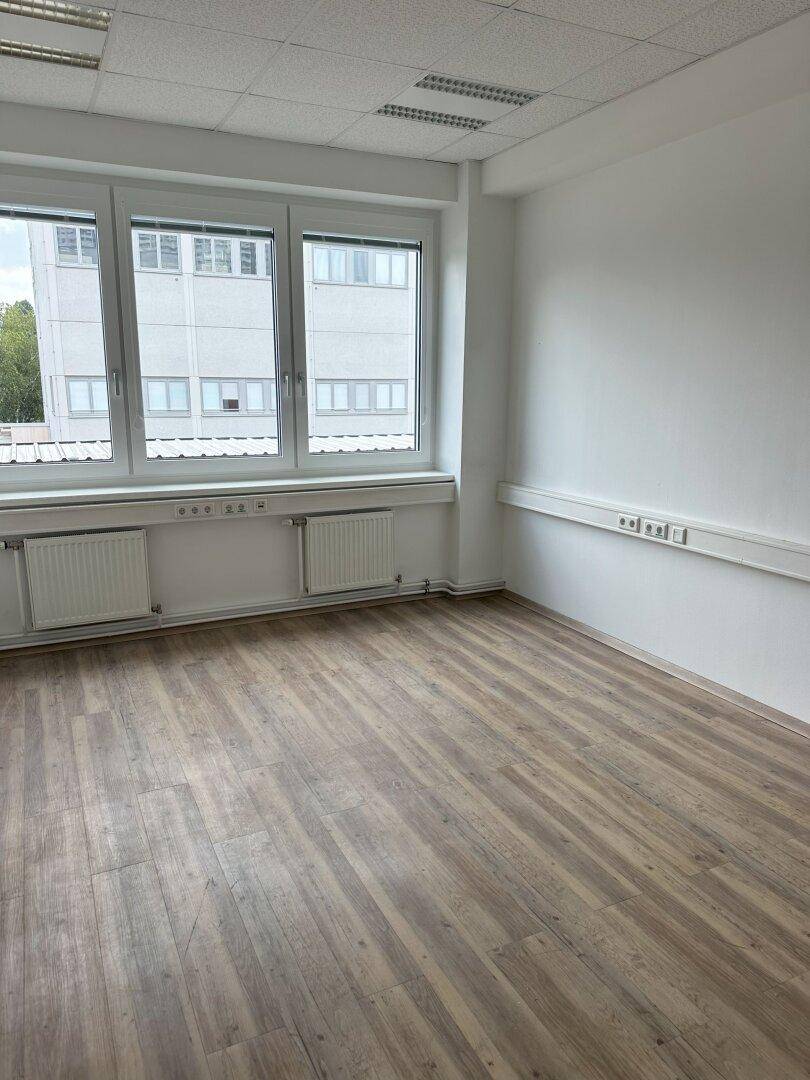 Helles, leeres Büro mit großen Fenstern, die einen Blick auf ein gegenüberliegendes Gebäude ermöglichen.