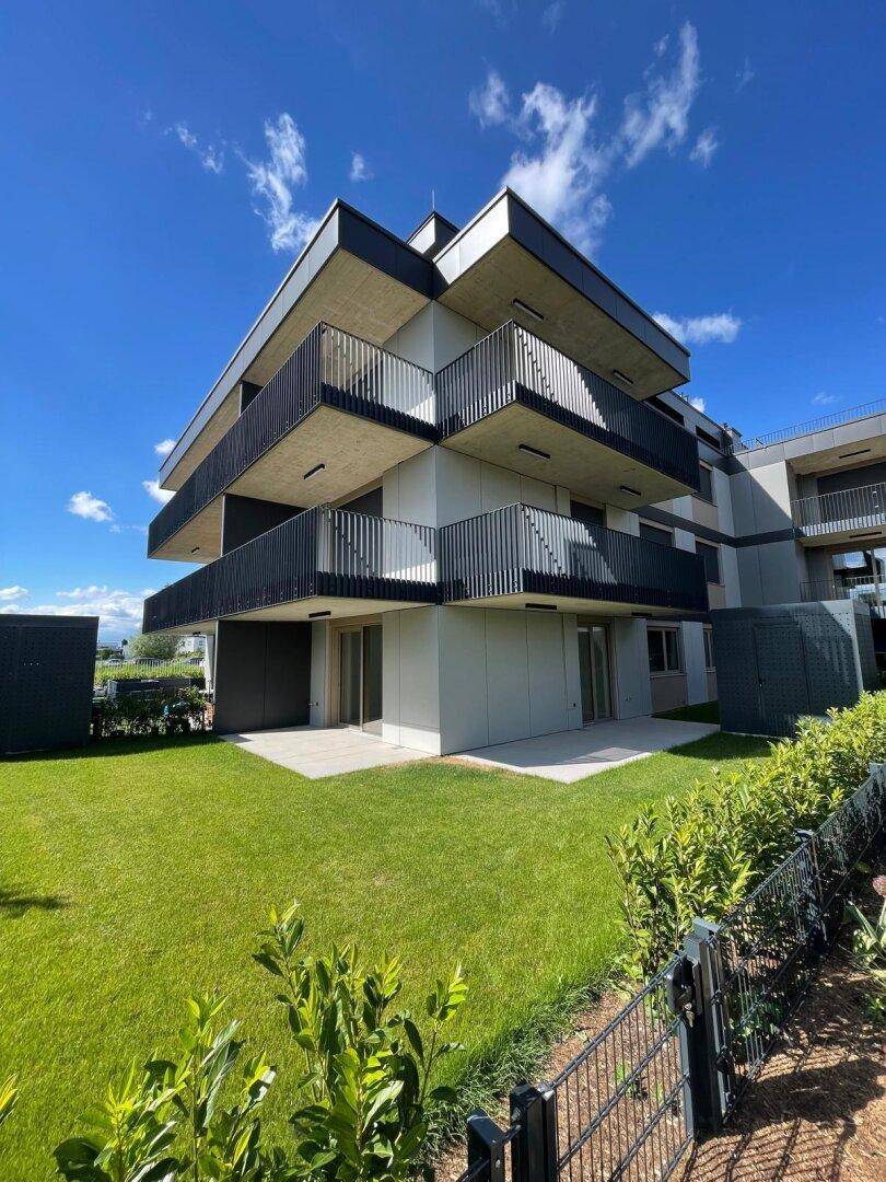 Modernes Mehrfamilienhaus mit mehreren Balkonen, einer Terrasse und gepflegtem Gartenbereich unter blauem Himmel.