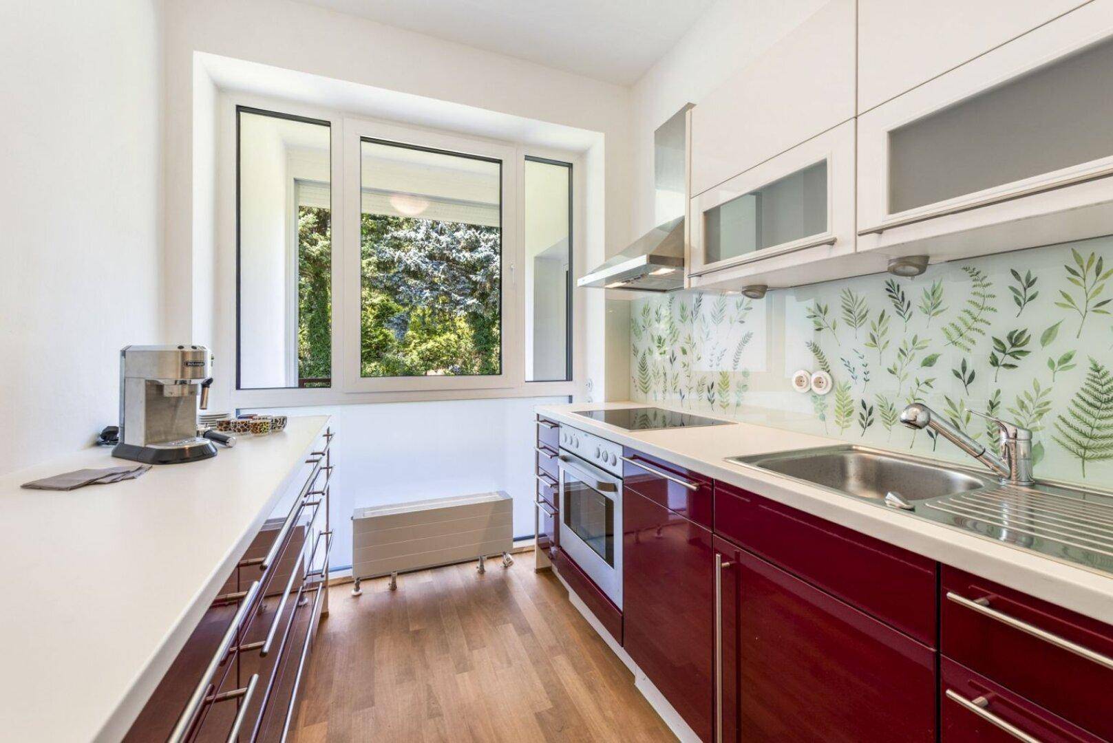 Moderne Küche mit roten Unterschränken, weißen Oberschränken und Blick ins Grüne durch das Fenster.