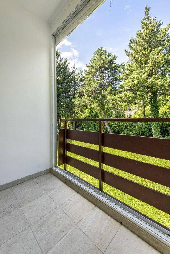 Sonniger Balkon mit Fliesenboden und Holzgeländer, umgeben von üppigem Grün und hohen Bäumen.