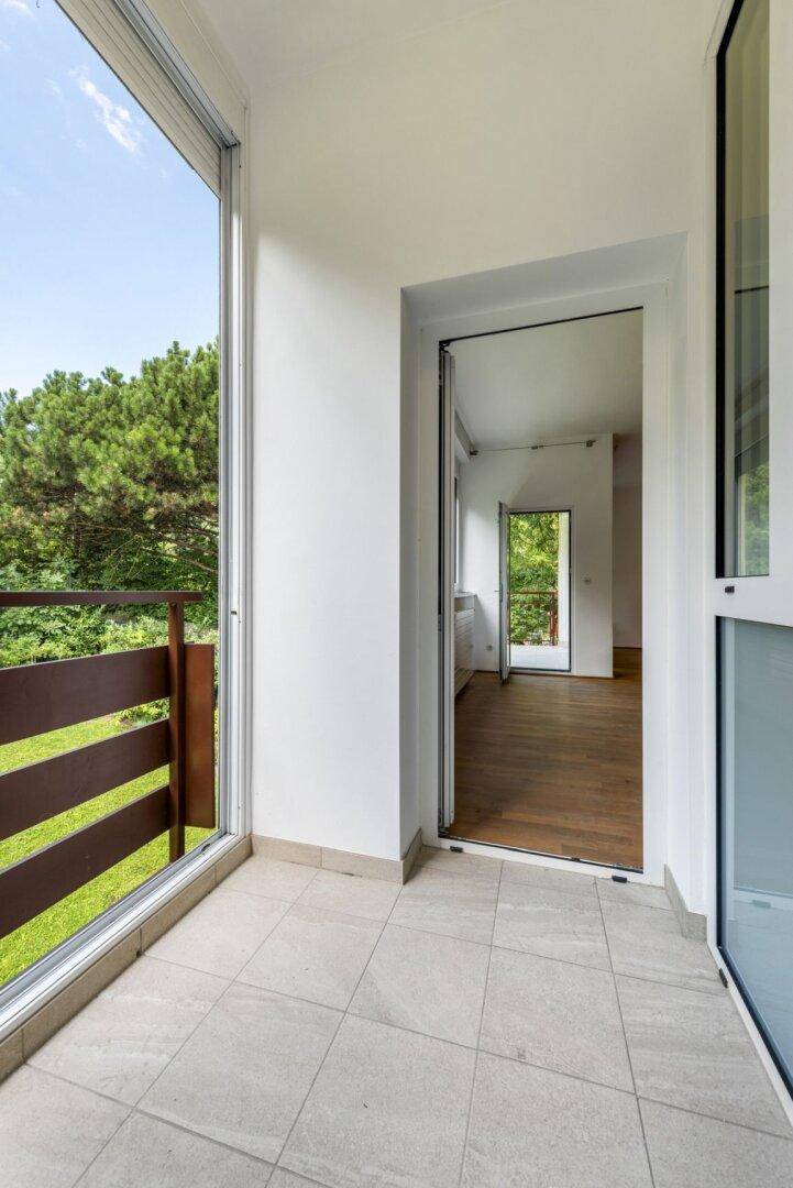 Heller Balkon mit Fliesenboden und Holzgeländer, der einen ruhigen Blick ins Grüne ermöglicht.