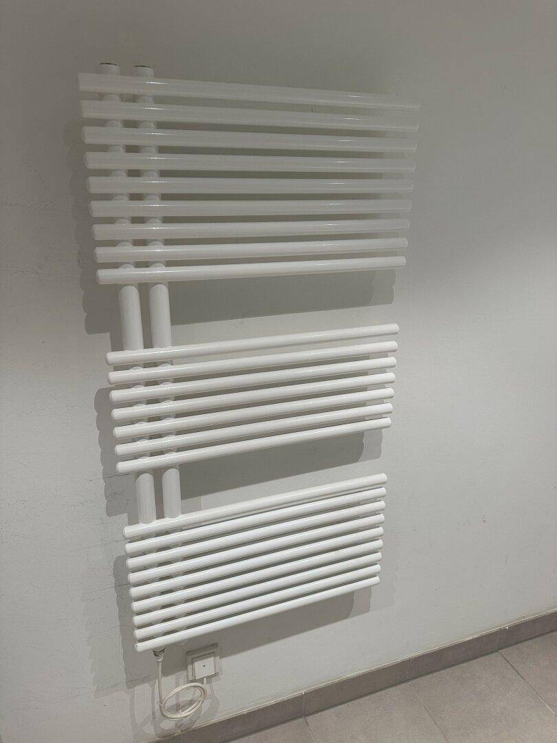 Weißer, moderner Handtuchheizkörper an einer hellgrauen Wand in einem Badezimmer mit Fliesenboden.