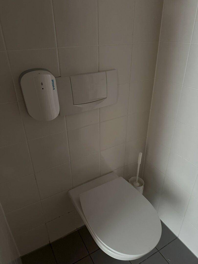Einfache Toilette mit Wand-WC, Fliesenboden und einem Spender an der Wand.