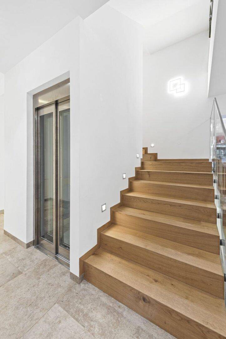Moderner Flur mit Holztreppe und integriertem Aufzug, hell beleuchtet und mit Fliesenboden ausgestattet.
