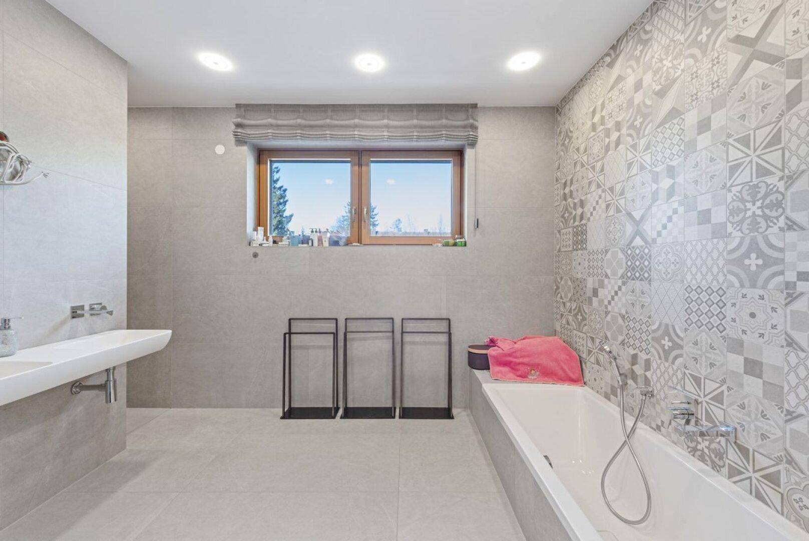 Modernes Badezimmer mit grauen Fliesen, Badewanne und Fenster mit Ausblick in die Natur.
