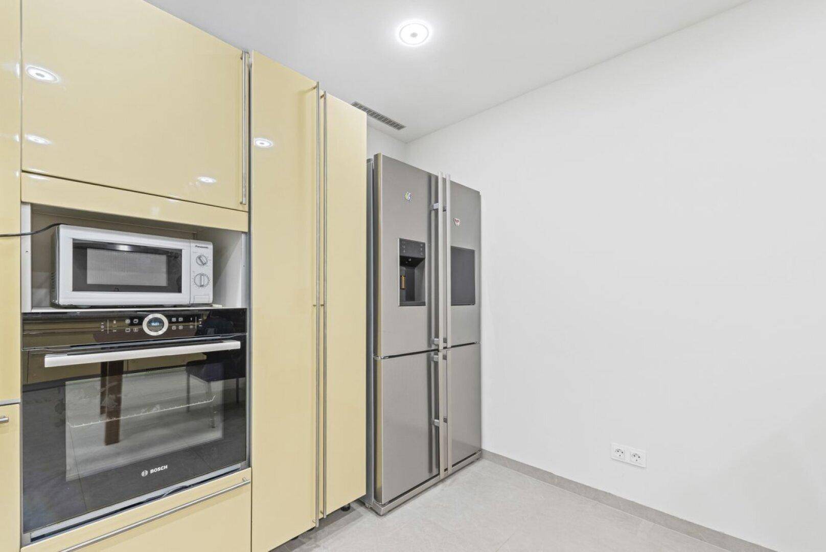 Moderne Küche mit beigen Hochglanzschränken, integriertem Backofen, Mikrowelle und großem Kühlschrank.