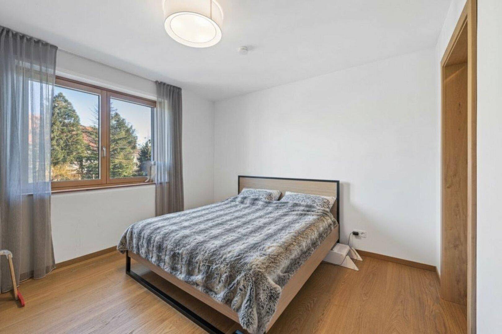 Gemütliches Schlafzimmer mit Doppelbett, großem Fenster und hellem Parkettboden, bietet eine ruhige Atmosphäre.