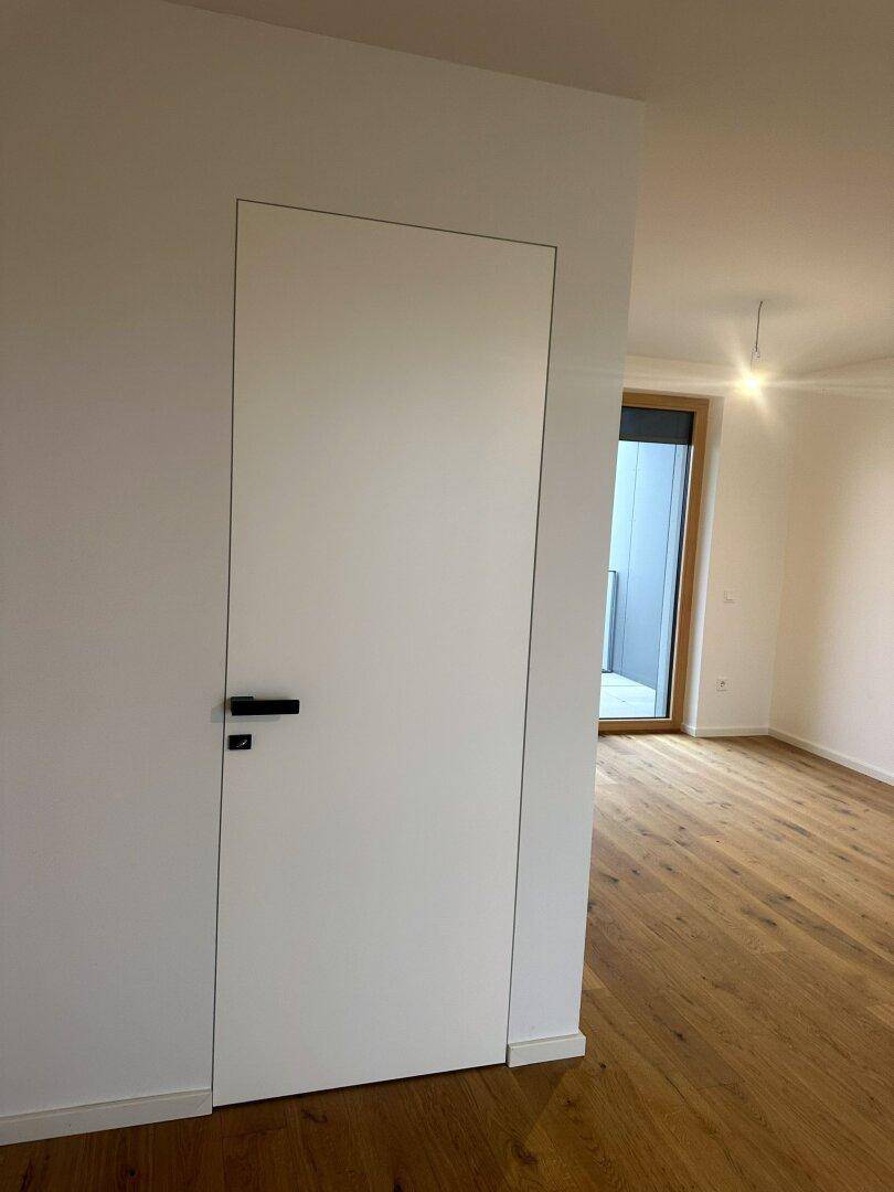 Moderner Innenraum mit einer rahmenlosen Tür und Holzboden, der zu einem weiteren Raum führt.