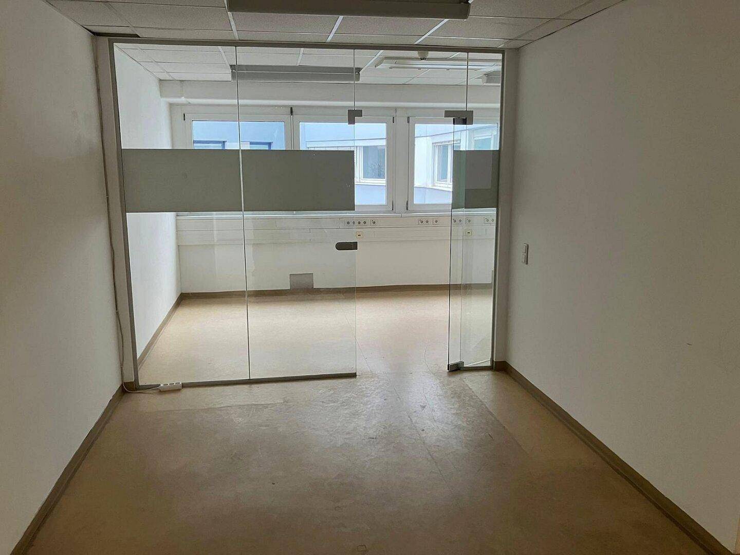 Leerer Raum mit einer Glaswand, die zu einem weiteren Bereich mit Fenstern führt und viel Licht hereinlässt.