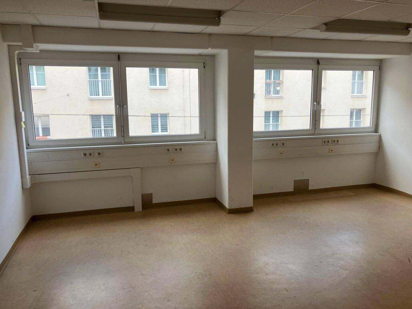 Leerer Büroraum mit großen Fenstern, die viel Tageslicht hereinlassen und einen Blick auf ein Gebäude bieten.