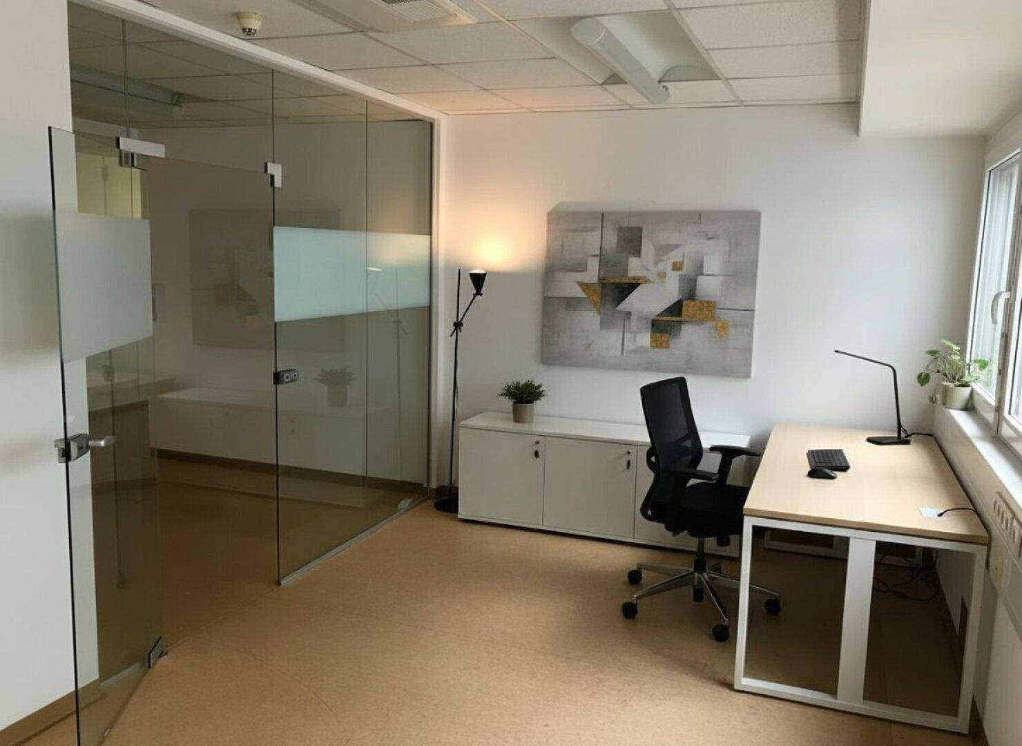Modernes Büro mit Glaswand, Schreibtisch, Bürostuhl und einem Schrank, dekoriert mit Kunst an der Wand.