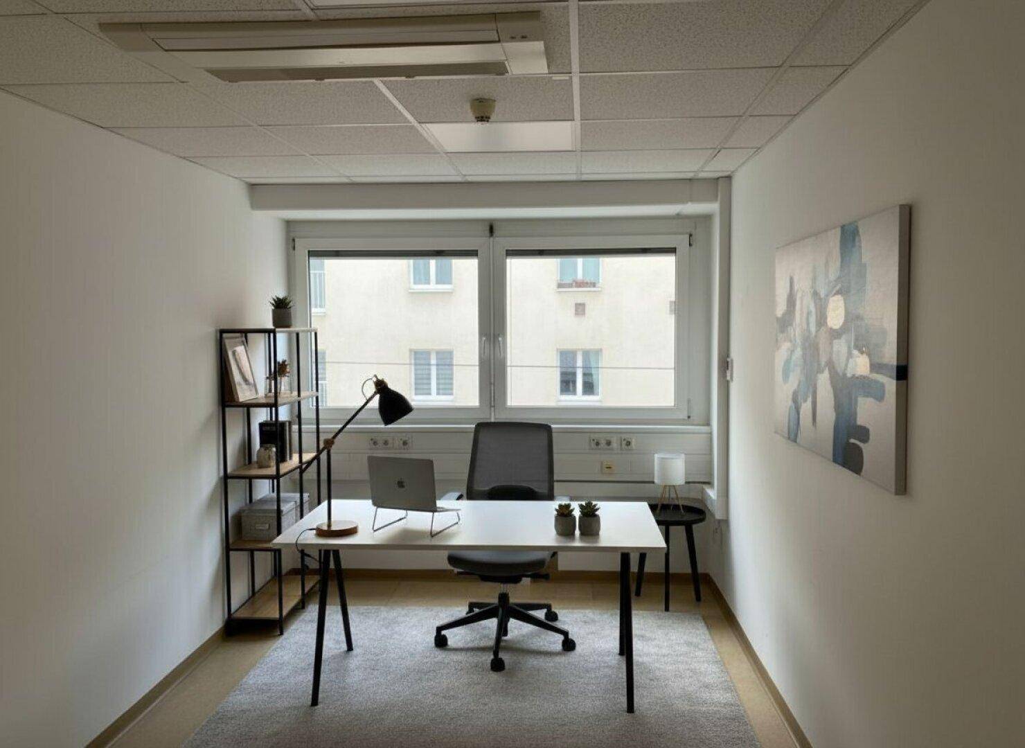 Kleines, helles Büro mit Schreibtisch, Bürostuhl, Laptop und einem Regal, mit Blick auf ein Gebäude.