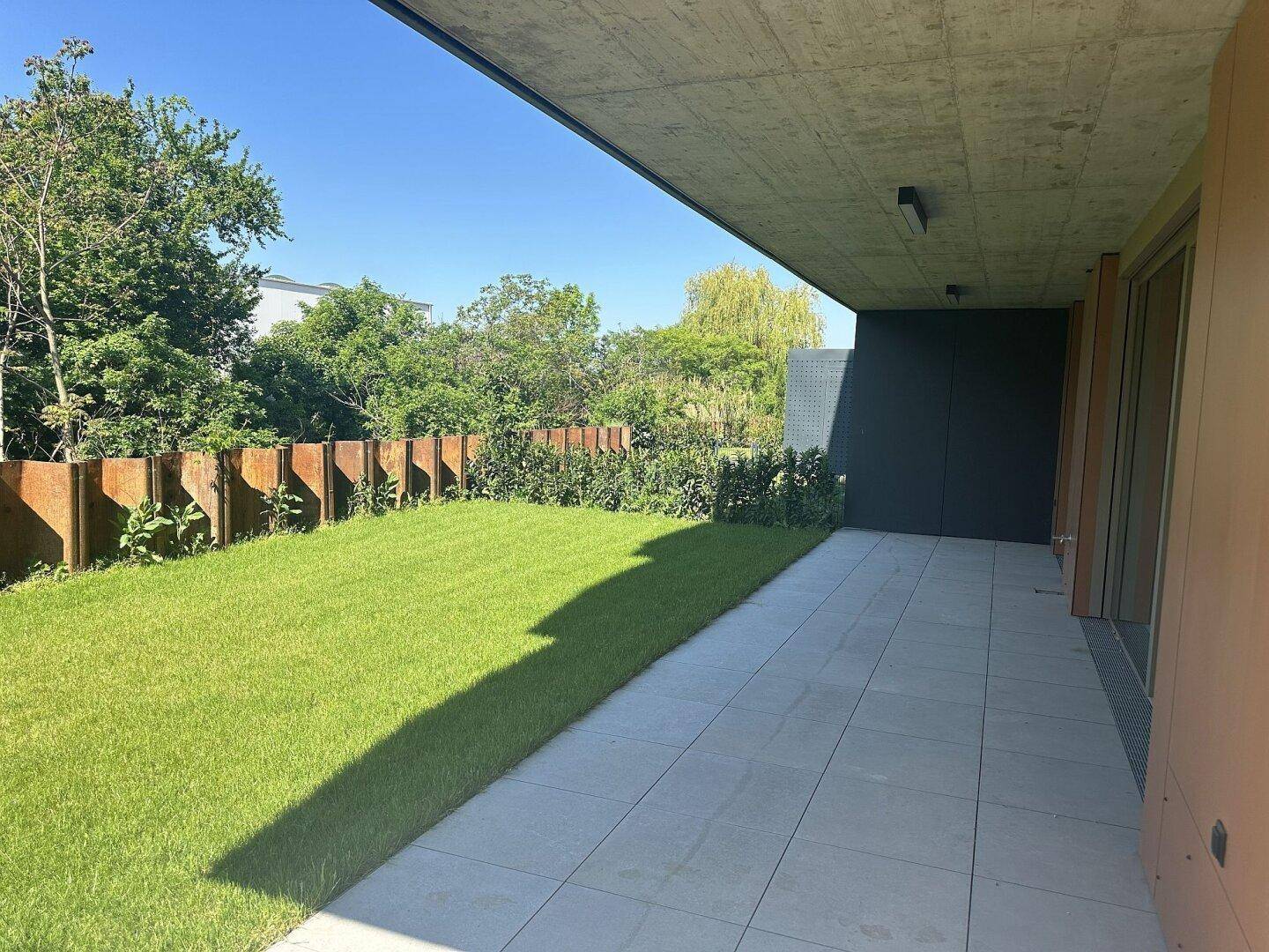 Privater Gartenbereich mit Rasenfläche und gefliester Terrasse unter einem überdachten Bereich.