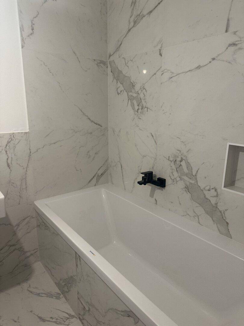 Elegantes Badezimmer mit integrierter Badewanne, Marmorfliesen und modernen schwarzen Armaturen.