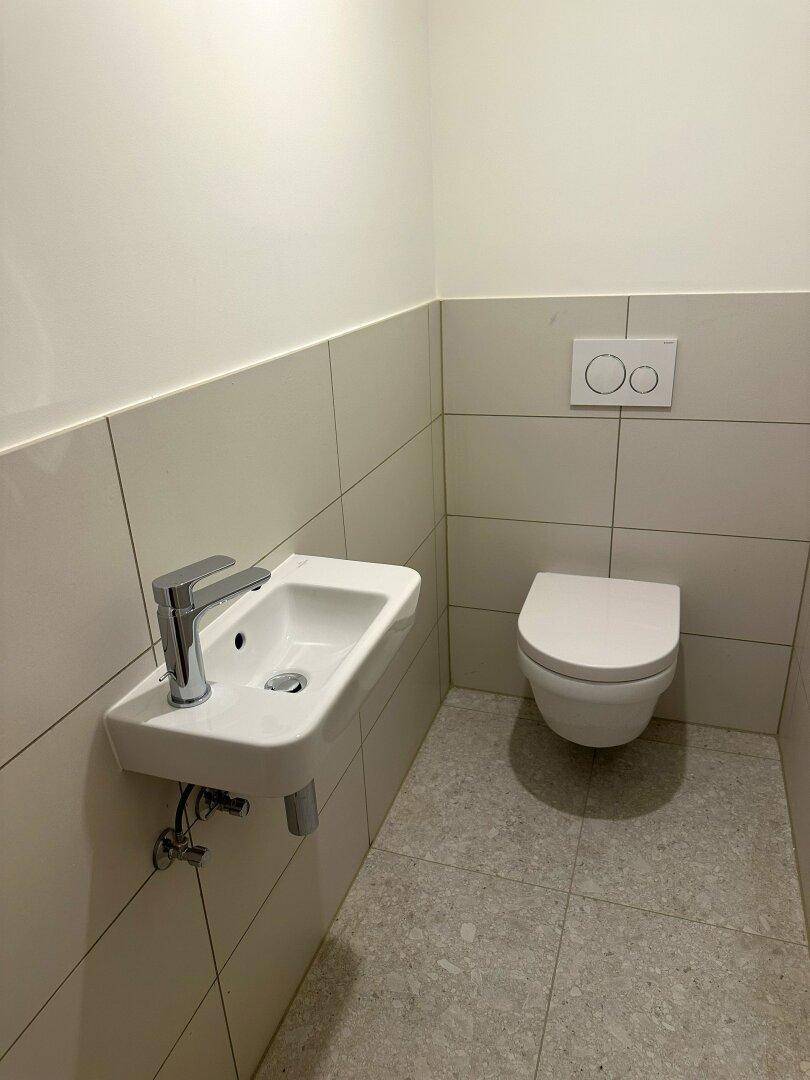 Modernes Gäste-WC mit Wand-WC und kleinem Waschbecken, in hellen Fliesen gehalten.