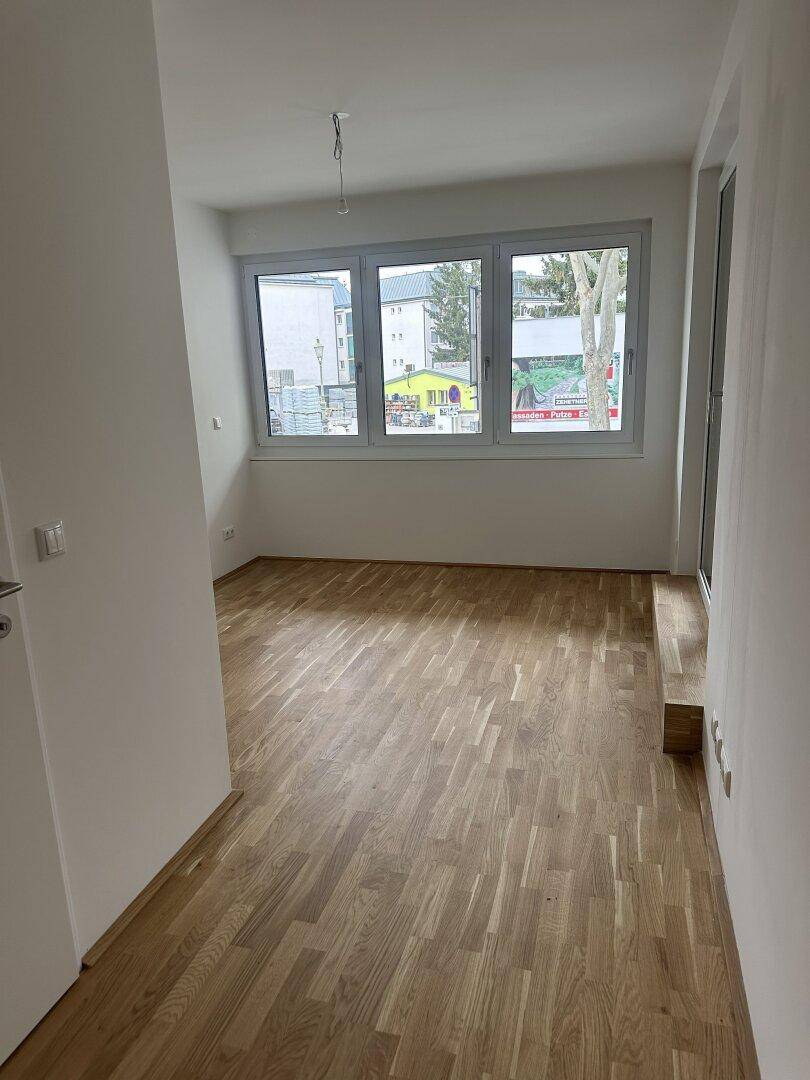 Leerer Raum mit Holzboden und großen Fenstern, die viel Tageslicht hereinlassen.