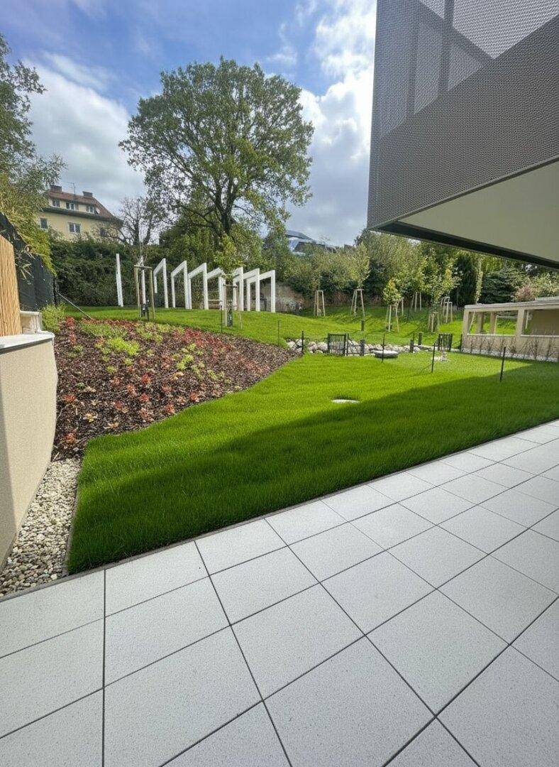 Gepflegter Gartenbereich mit Rasenfläche und moderner Terrasse, umgeben von Bäumen und Sträuchern.