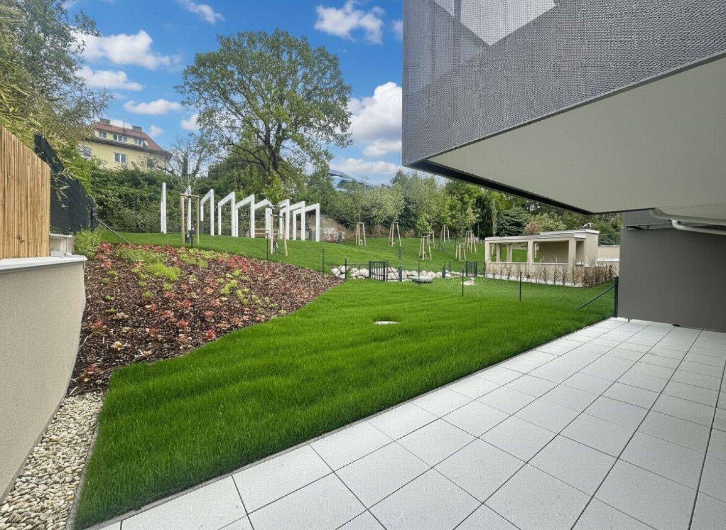 Gepflegter Gartenbereich mit Rasenfläche und moderner Terrasse, umgeben von Bäumen und Sträuchern.