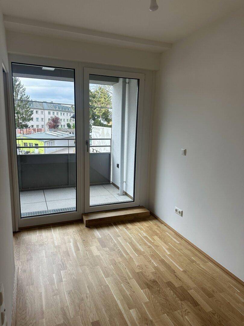 Heller, leerer Raum mit Holzboden und großen Glasfenstern, die zu einem Balkon führen.