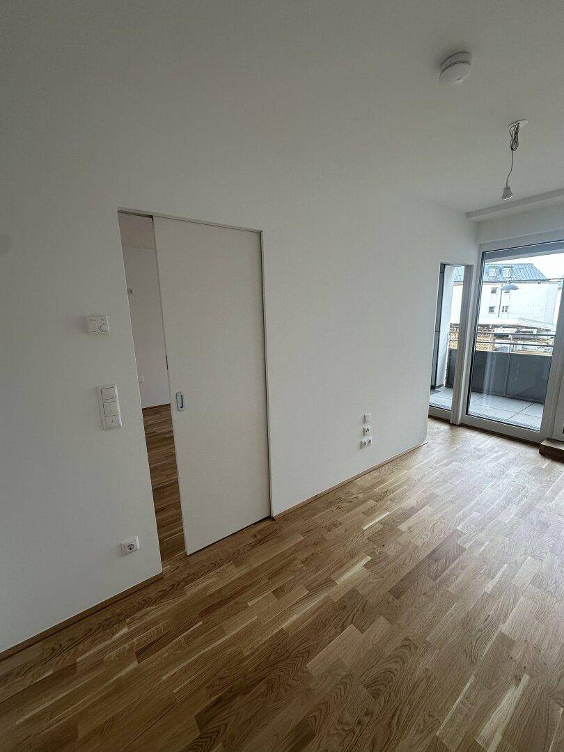 Leerer Raum mit Holzboden, weißen Wänden, Schiebetür und Zugang zu einem Balkon.