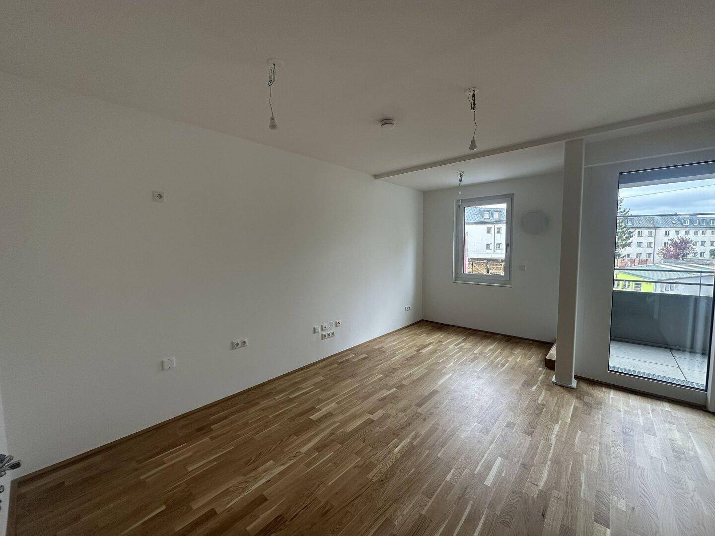 Geräumiger, leerer Raum mit Holzboden, weißen Wänden und Fenster, mit Balkonzugang.
