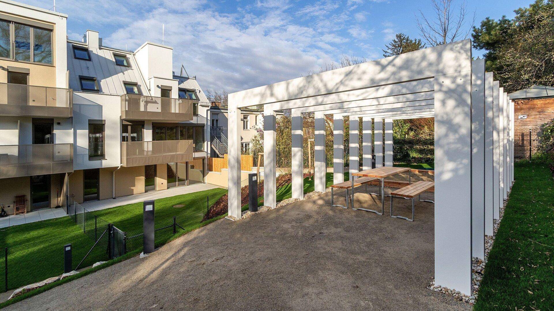 Moderner Außenbereich mit Pergola, Picknicktischen und gepflegter Rasenfläche neben dem Wohngebäude.