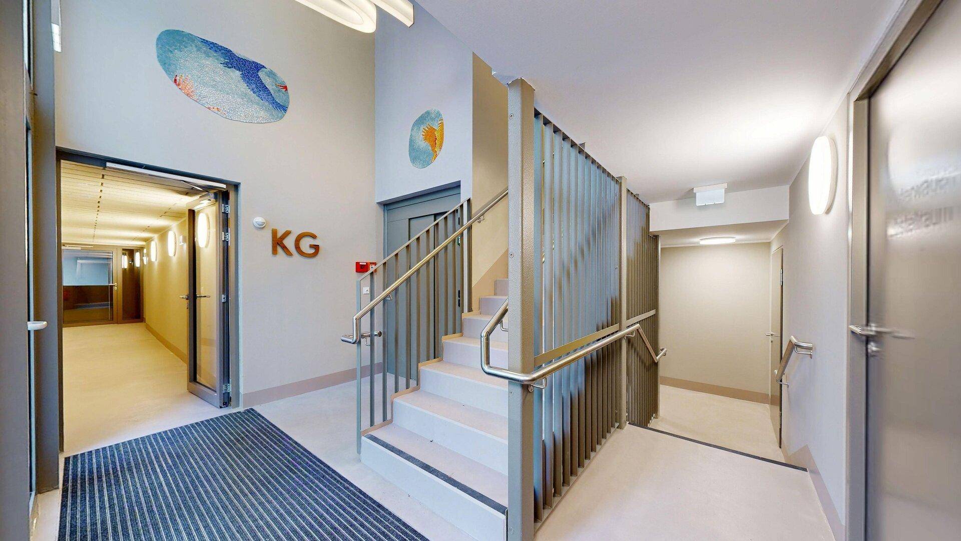 Heller Eingangsbereich mit Treppe, Rampe und Zugang zu weiteren Fluren im modernen Gebäude.