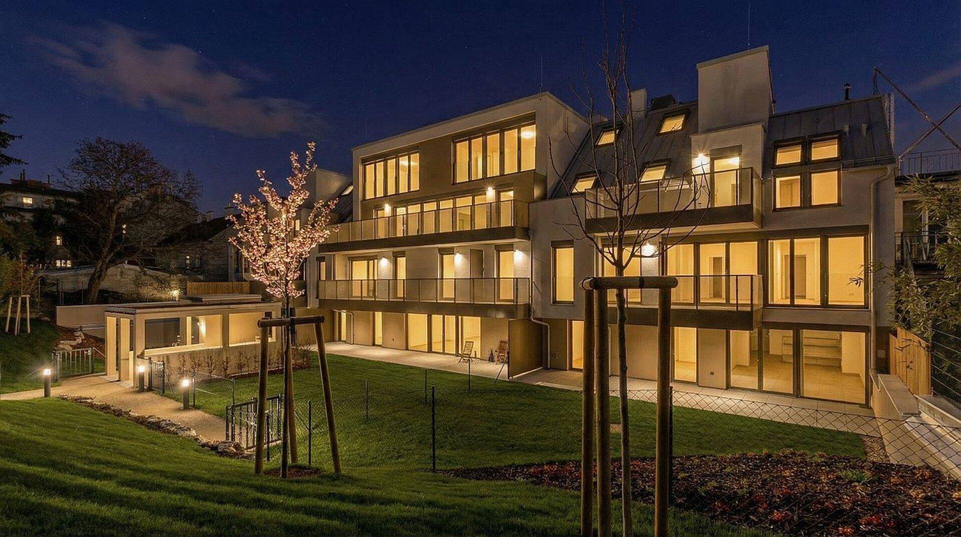 Modernes Mehrfamilienhaus bei Nacht mit beleuchteten Fenstern und gepflegtem Gartenbereich.