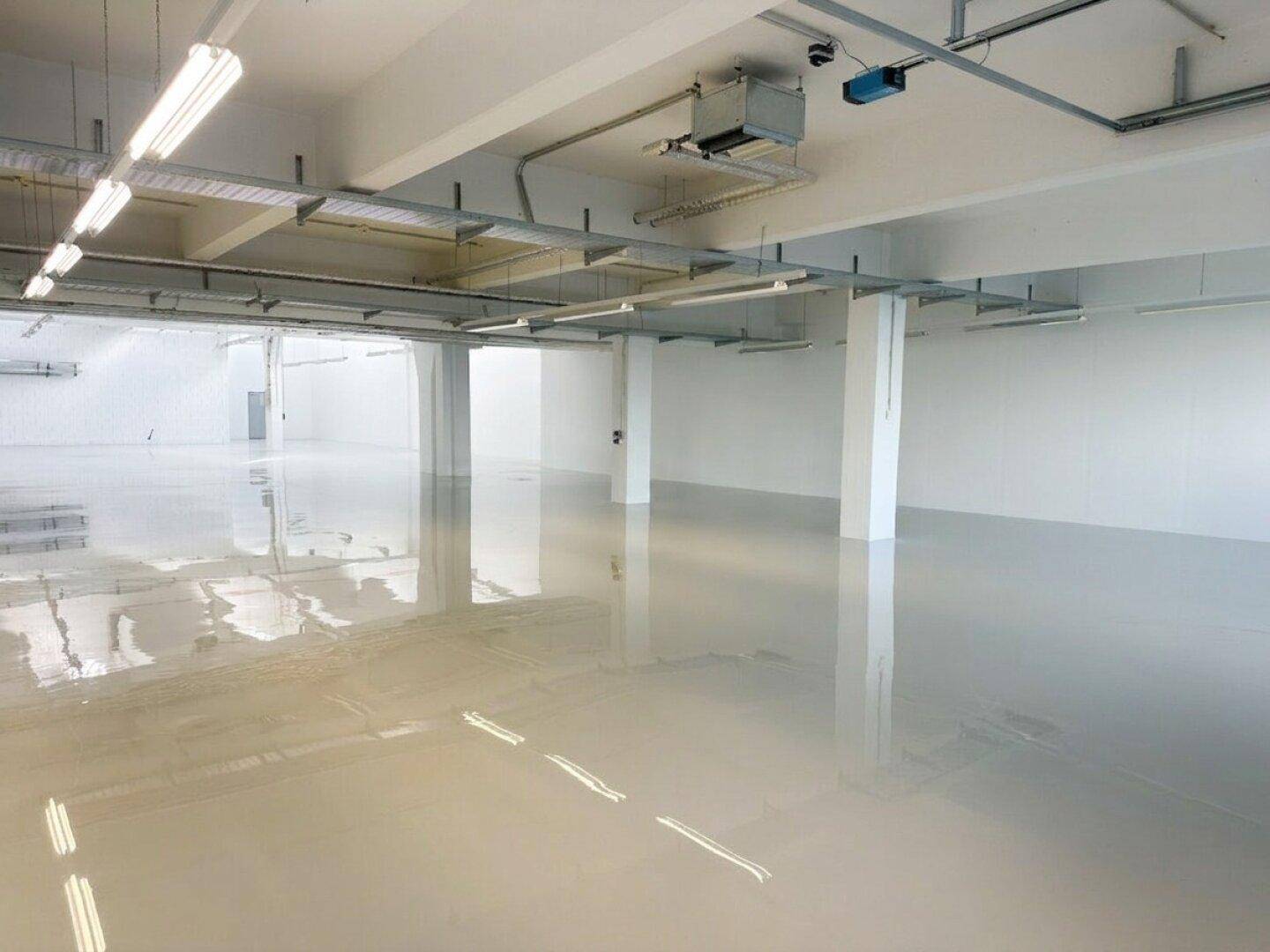 Großer, leerer Industrieraum mit glänzendem Boden, Säulen und sichtbaren Deckeninstallationen.