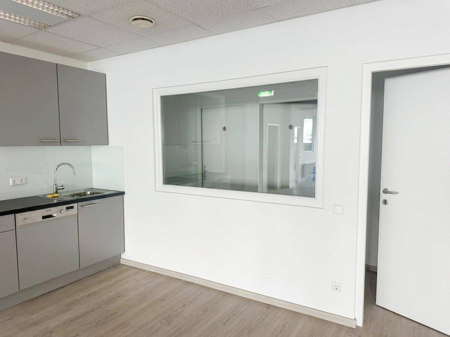 Kleine Küchenzeile in einem Büroraum mit Spüle, Geschirrspüler und grauen Schränken.