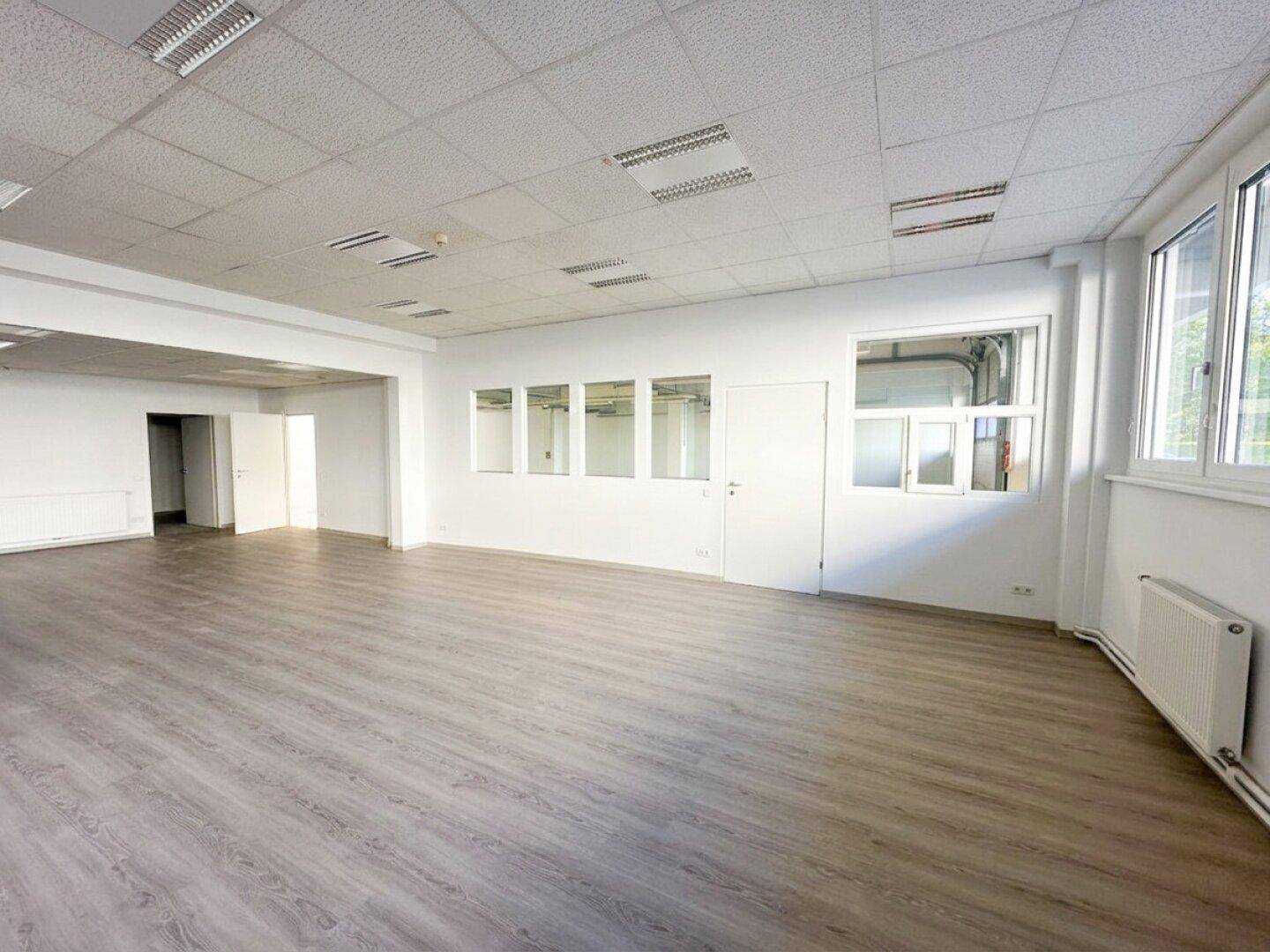Großer Büroraum mit Laminatboden, Fenstern und einer Tür zu einem weiteren Bereich.