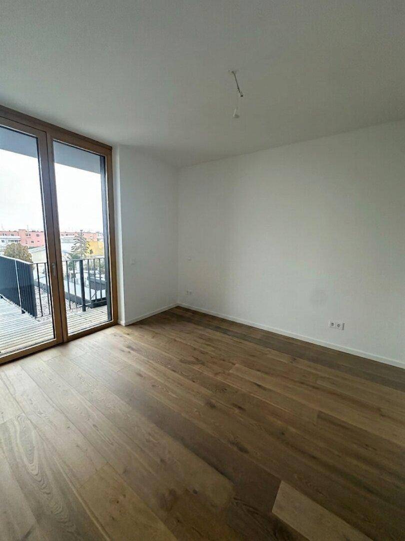 Heller, leerer Raum mit Holzboden und großen Fenstern, die viel Tageslicht hereinlassen.