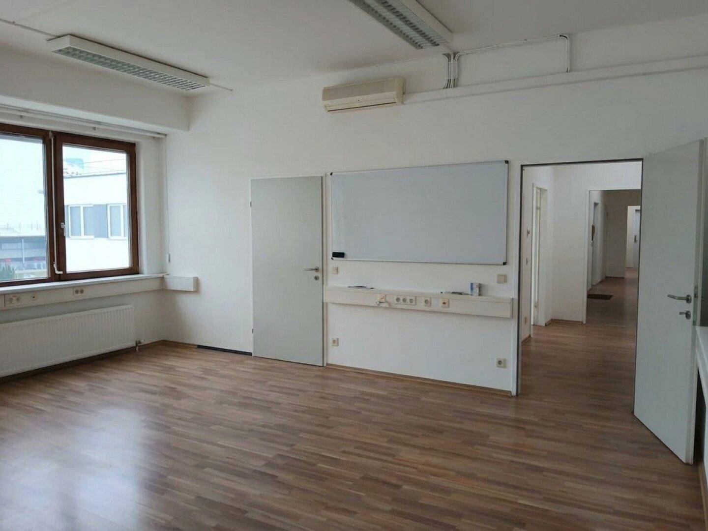 Leerer Büroraum mit Laminatboden, Klimaanlage, Whiteboard und Zugang zu weiteren Räumen.