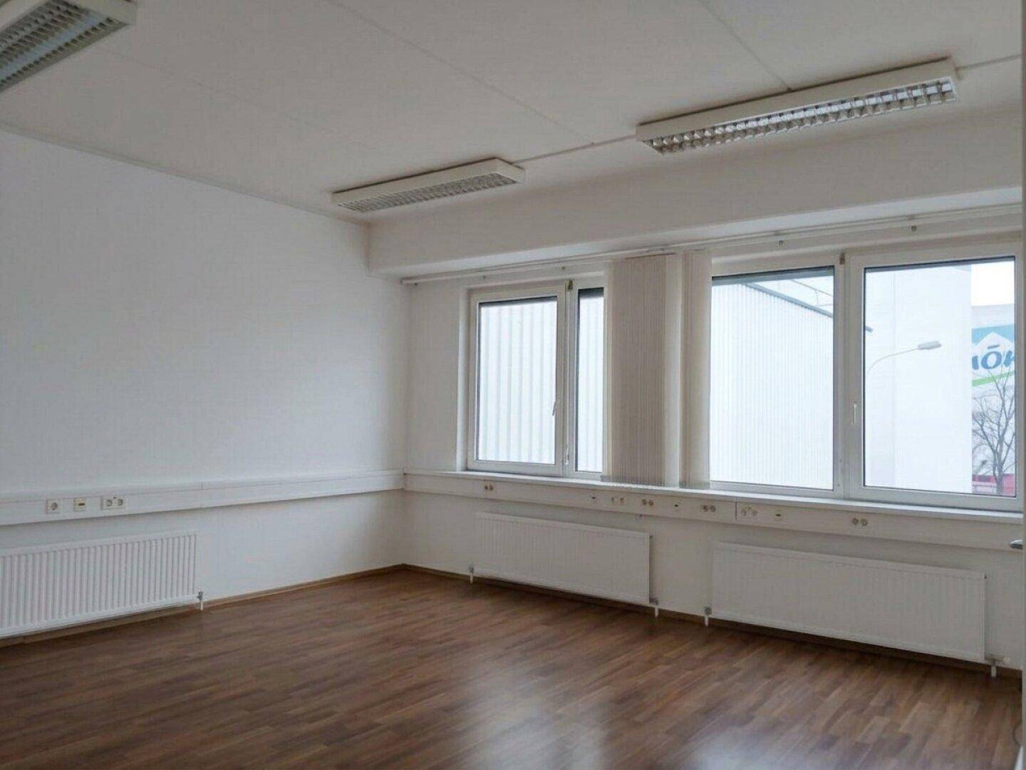 Geräumiger Büroraum mit Laminatboden, großen Fenstern und Deckenleuchten für gute Beleuchtung.
