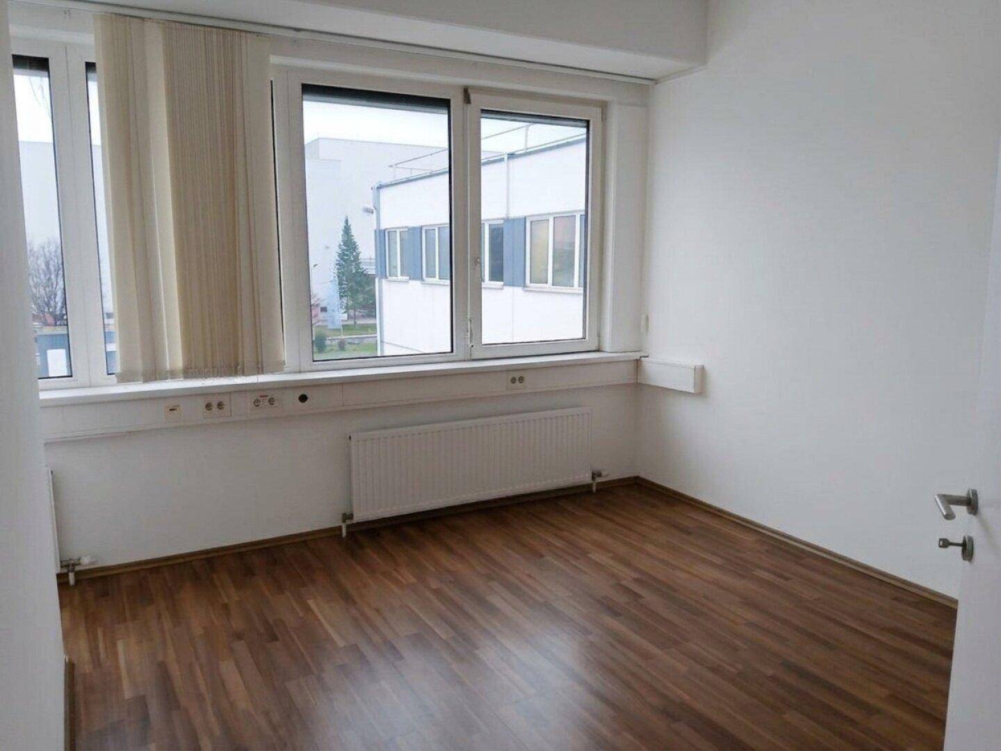 Heller Büroraum mit großen Fenstern, Laminatboden und Heizkörpern unter den Fenstern.
