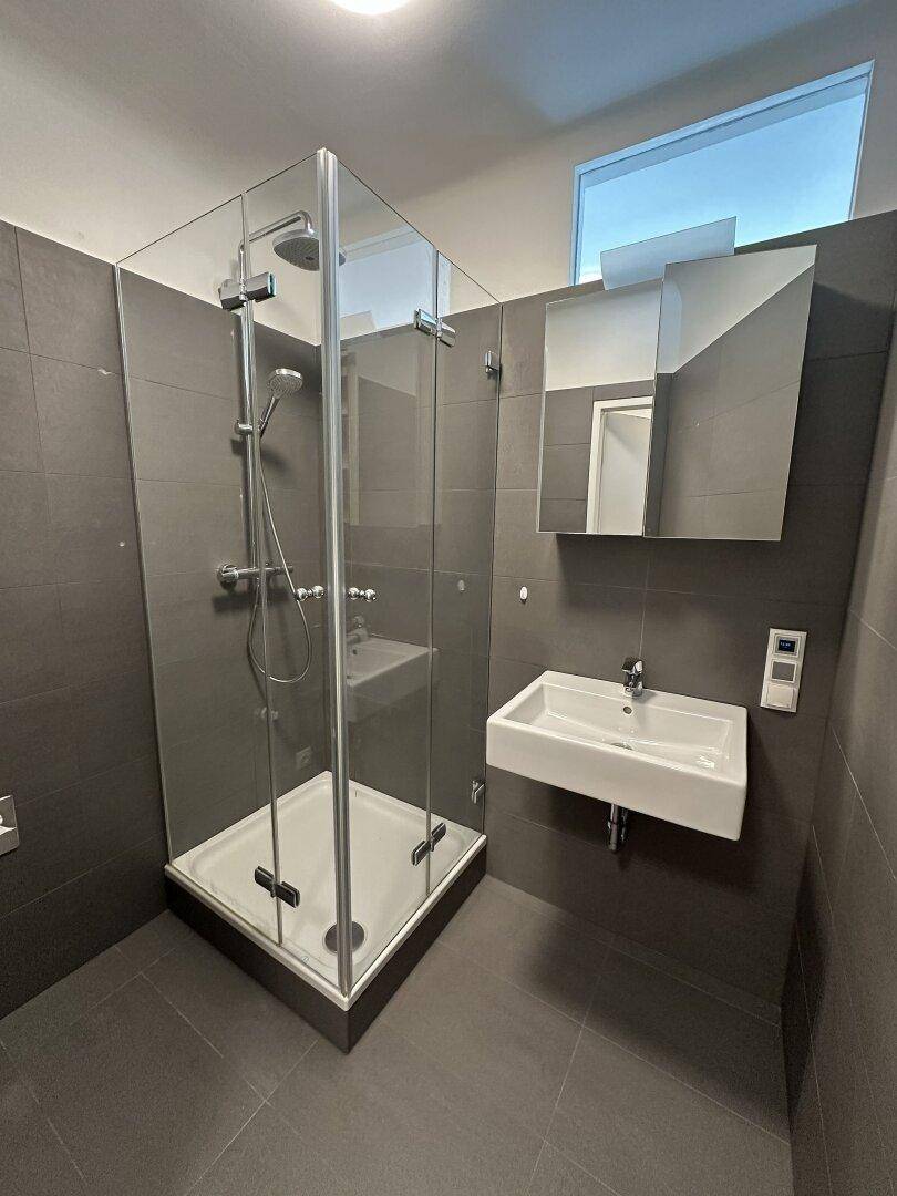 Modernes Badezimmer mit großer Glasdusche, Wandwaschbecken und grauen Fliesen.