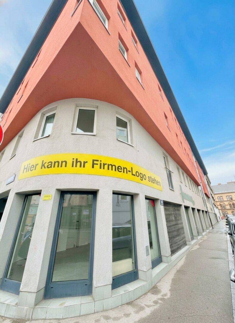 Eckgebäude mit moderner Architektur, großen Schaufenstern im Erdgeschoss und orangefarbenen Fassadenelementen.