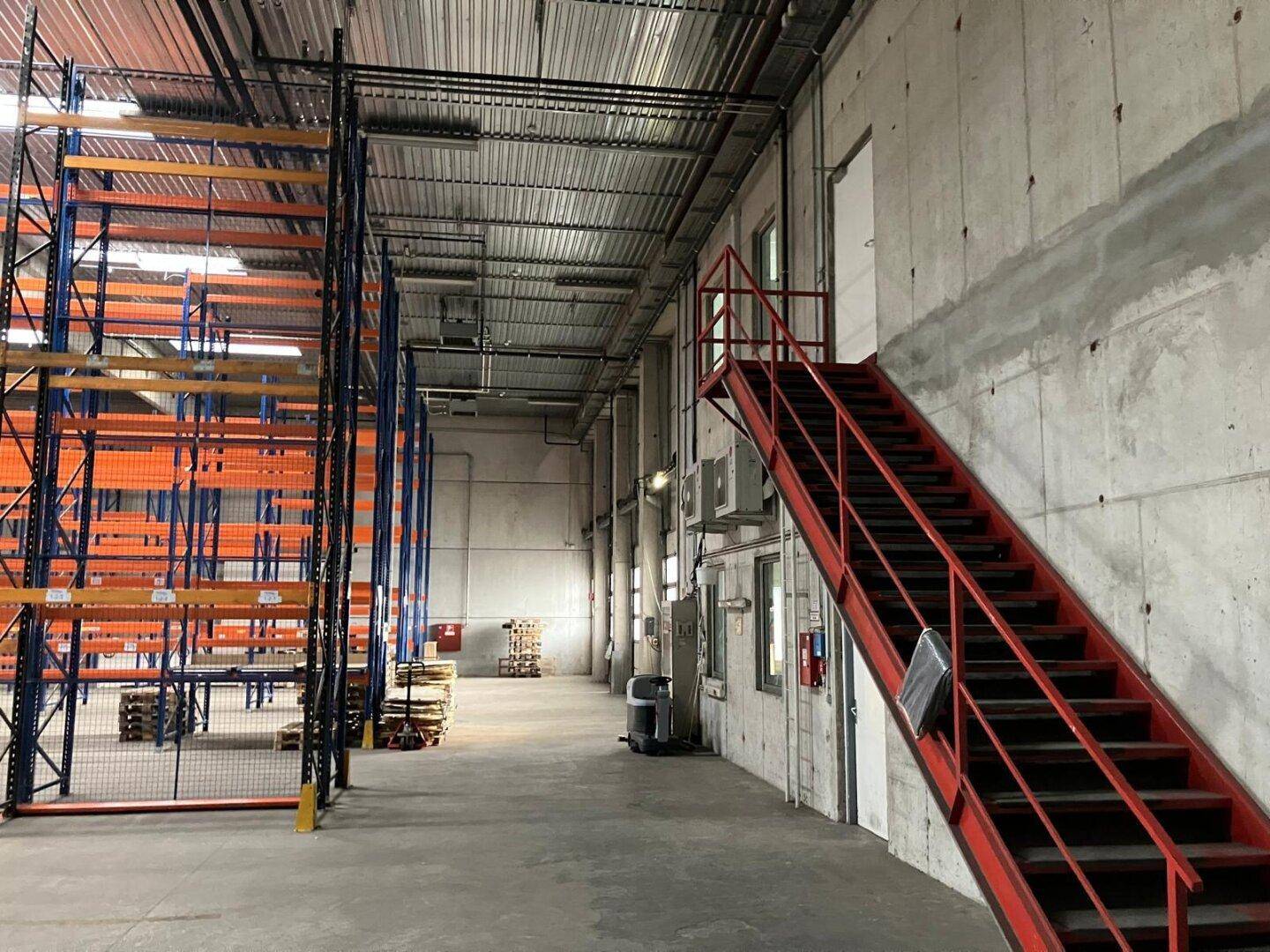Lagerhalle mit roter Metalltreppe zu einer oberen Ebene, Betonwänden und Regalsystemen.
