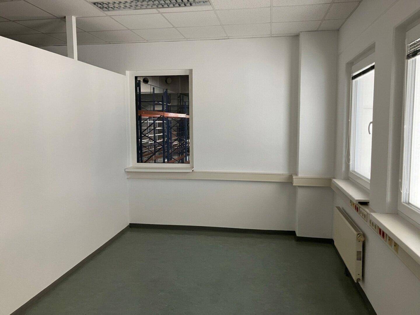 Helles Büro mit weißen Wänden, grauem Boden und Fensterblick in die Lagerhalle.
