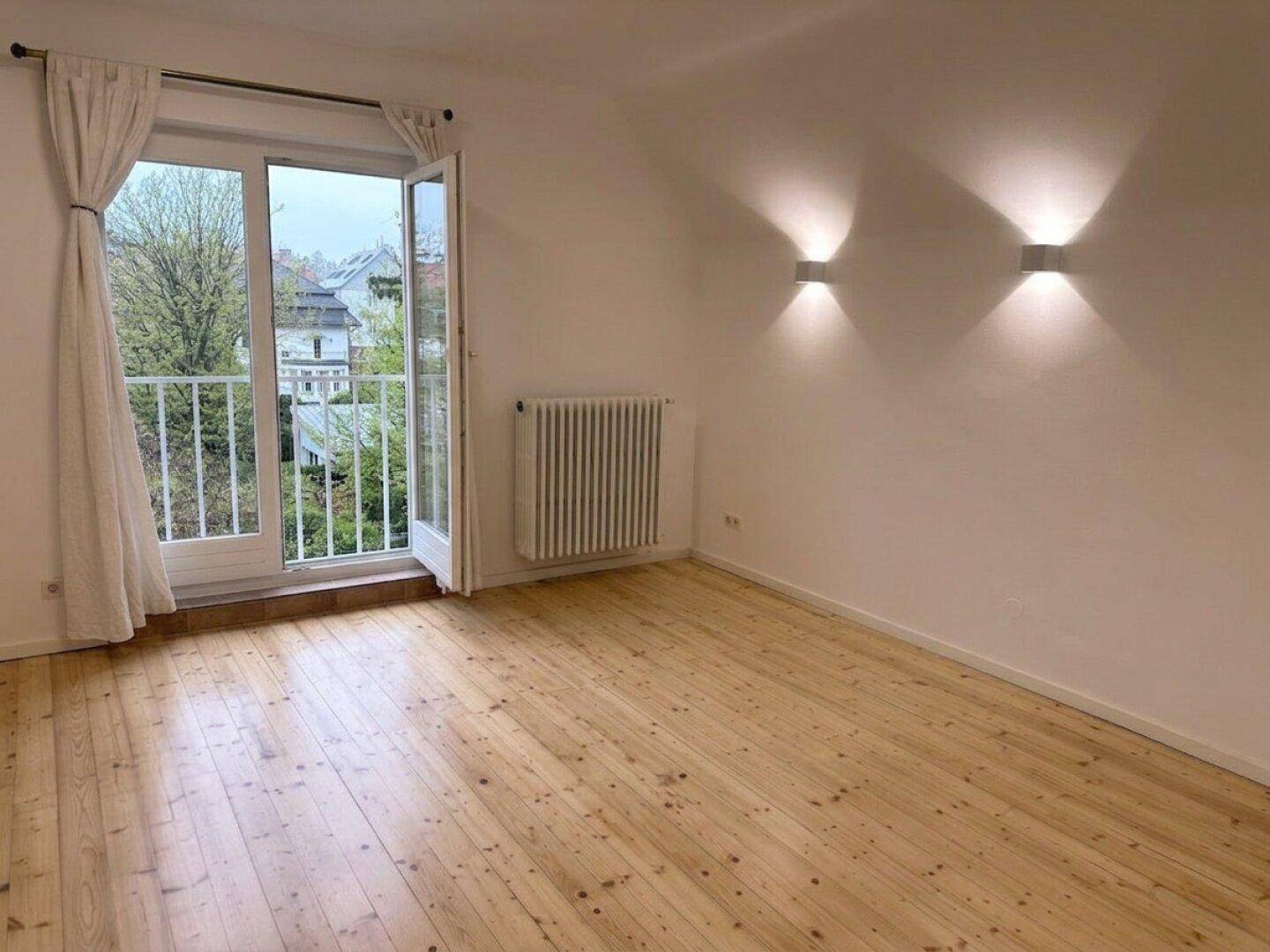 Lichtdurchfluteter Raum mit Holzboden und Blick auf die Umgebung durch die Balkontür.