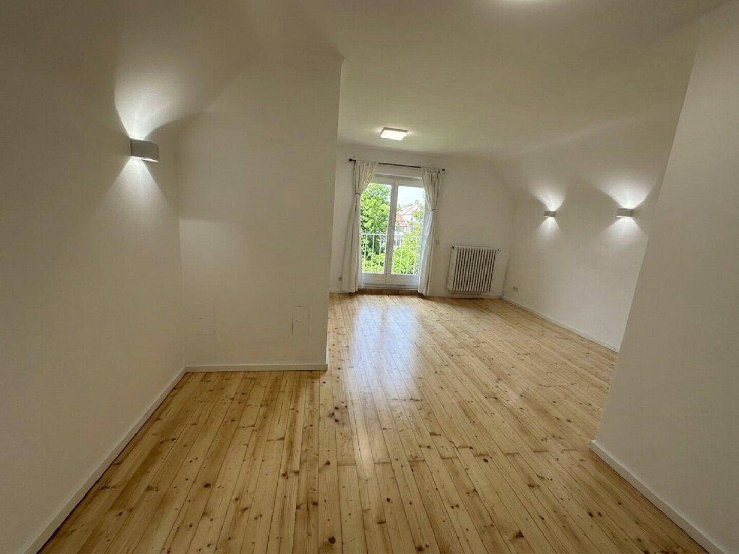 Geräumiger Wohnbereich mit hellem Holzboden und Zugang zu einem französischen Balkon.