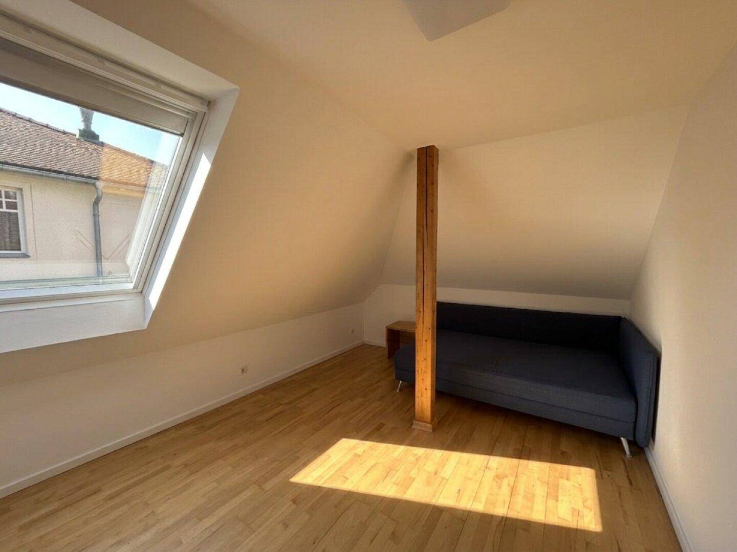 Helles Zimmer im Dachgeschoss mit Dachfenster, Holzbalken und Schlafsofa.