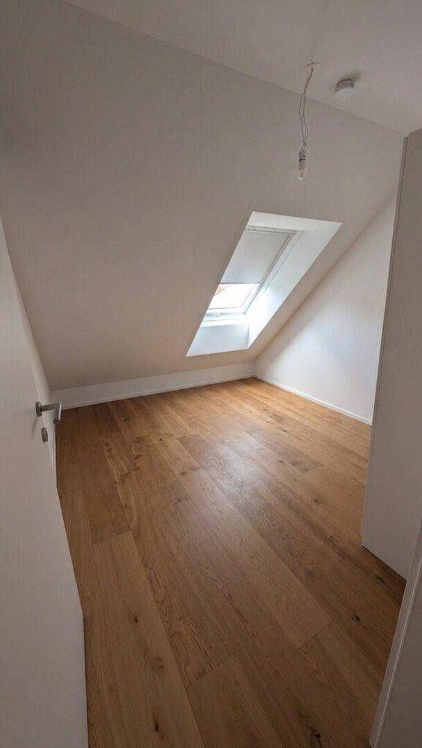 Helles Dachzimmer mit schrägen Wänden, Holzboden und einem großen Dachfenster, das viel Tageslicht spendet.