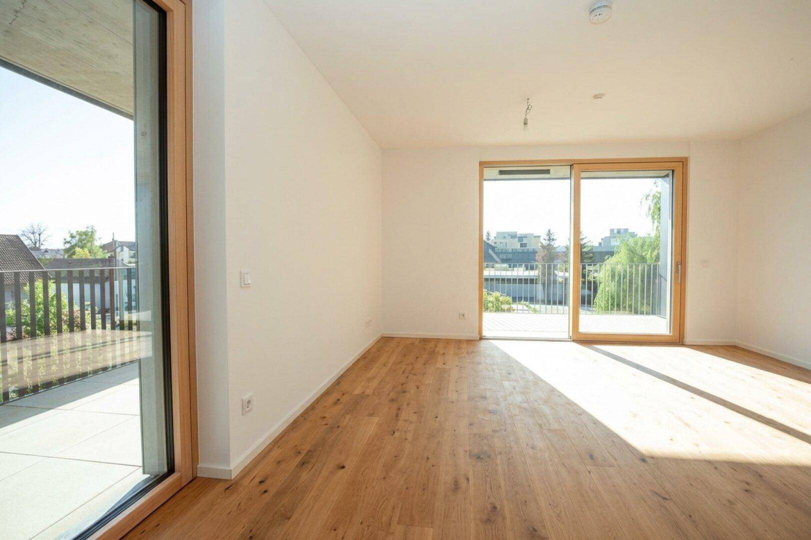 Heller, leerer Raum mit Holzboden und großen Schiebetüren, die auf einen sonnigen Balkon führen.