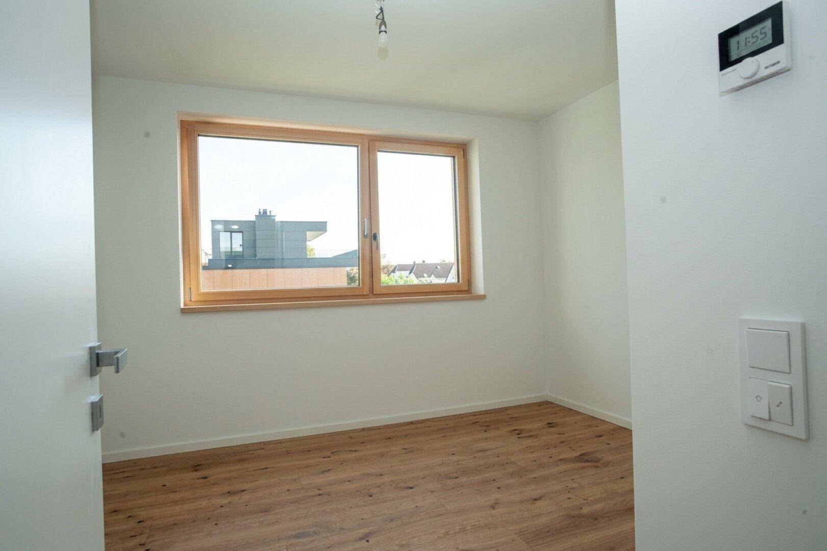 Leeres Schlafzimmer mit Holzboden, weiß gestrichenen Wänden und einem Fenster mit Blick nach draußen.