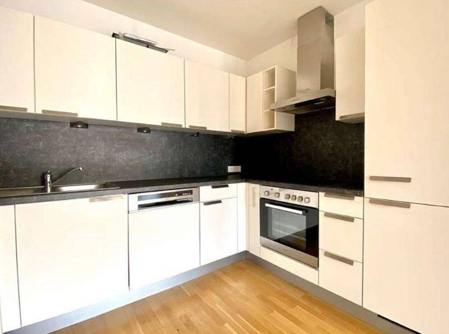 Moderne Einbauküche mit weißen Fronten, dunkler Arbeitsplatte und integrierten Geräten auf Holzparkett.